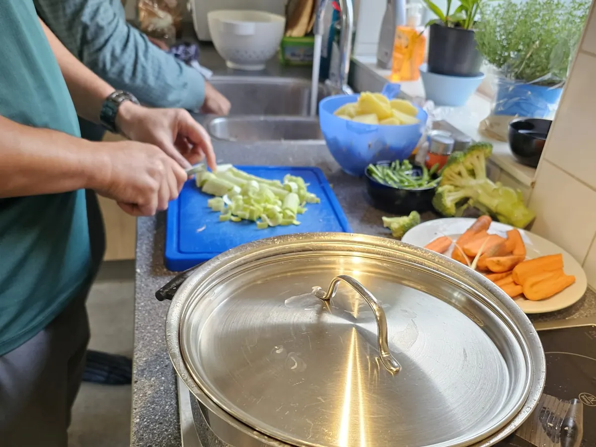 Handen snijden prei op een blauwe snijplank naast een grote pan, met wortels en broccoli klaar op het aanrecht.