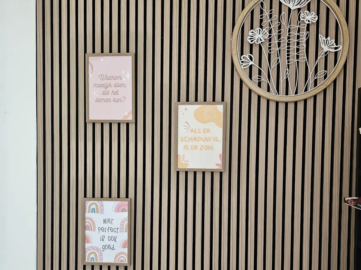 Een wand met houten latten in de huiskamer, met drie ingelijste posters en een ring met witte bloemen.