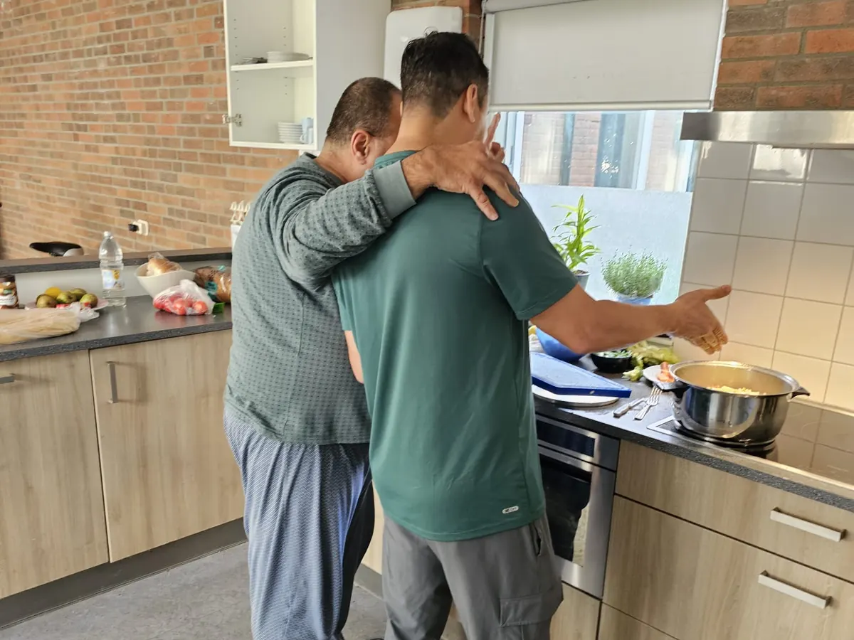 In de keuken slaat de een zijn arm om de schouder van de ander, terwijl die naar de pan op het fornuis wijst.