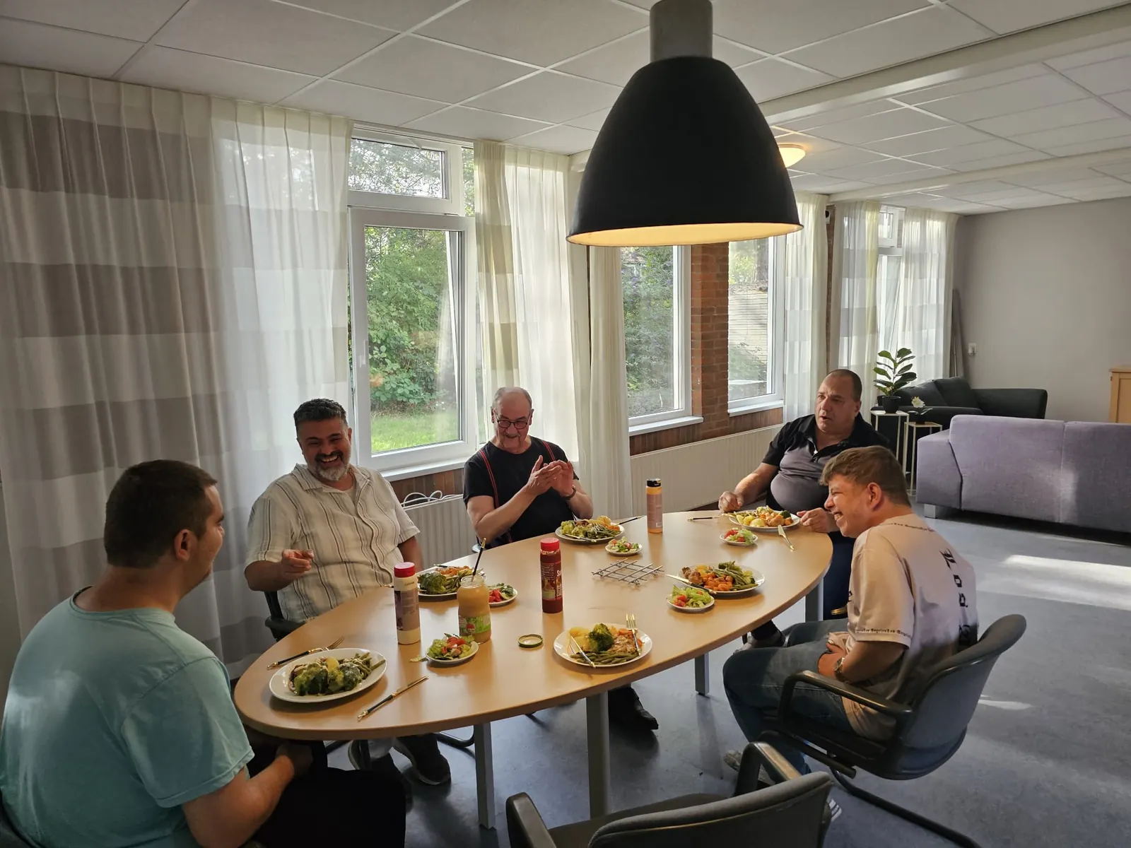 Vijf mannen aan een ovale tafel in de huiskamer, samen aan het eten en aan het lachen.