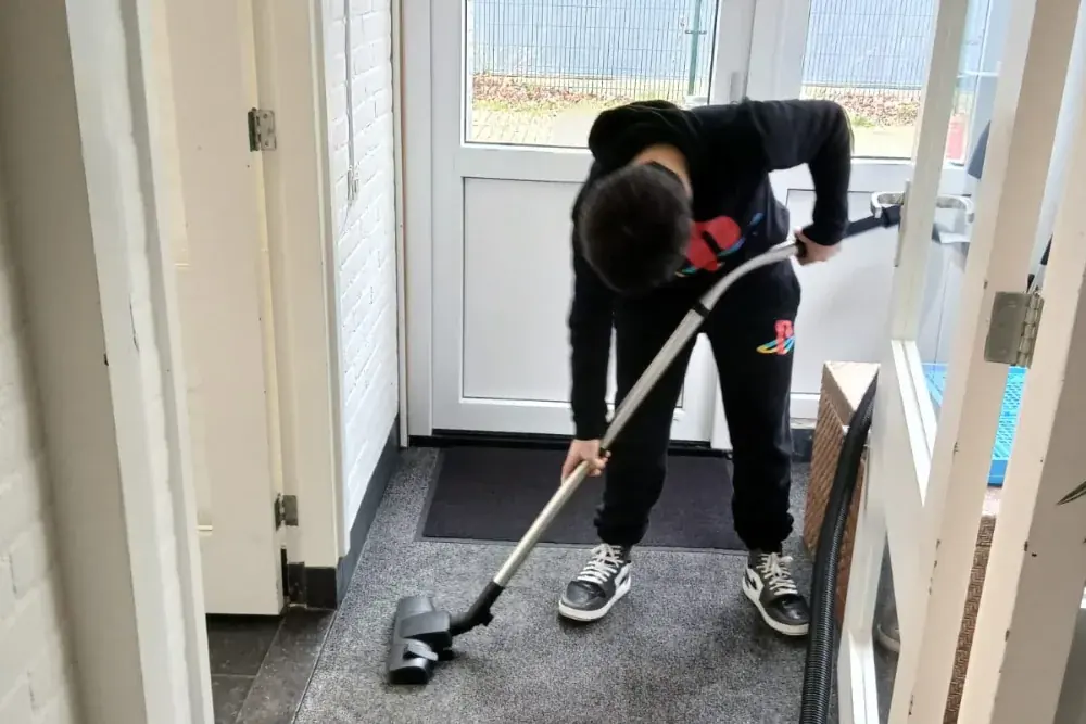 Iemand stofzuigt de mat bij de voordeur, met de rug naar de camera.
